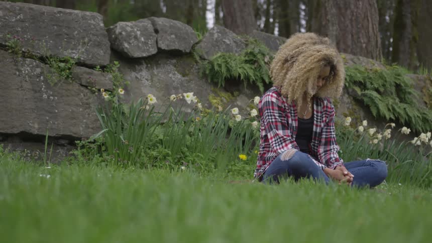 A young woman meditates while sitting in the grass in front of a stone wall