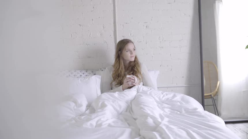 A woman drinks from a white mug while sitting in bed with a white blanket
