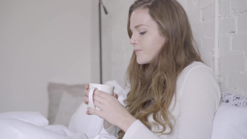 A woman is sitting in bed drinking from a mug