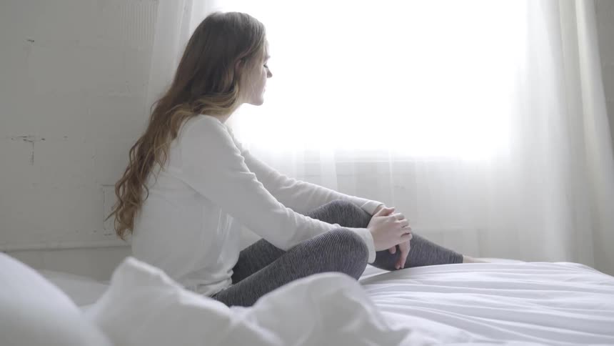 Woman wearing white long sleeve t shirt sits down on bed and looks out the window in the daytime