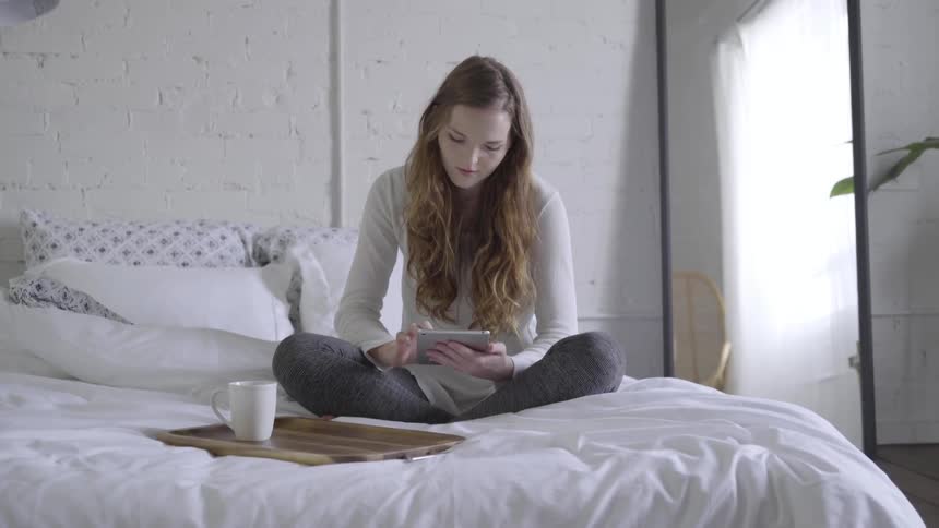 A young woman with long cascading brown hair sits on her bed in a well lit room with a tablet computer in her hands and a small tray with a coffee cup on it