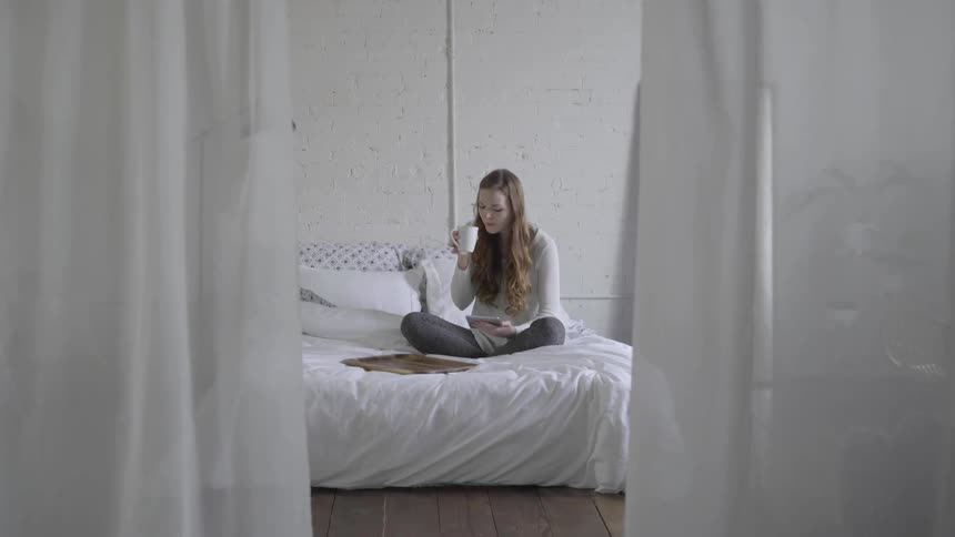 A young female sips a brew while sitting on her bed