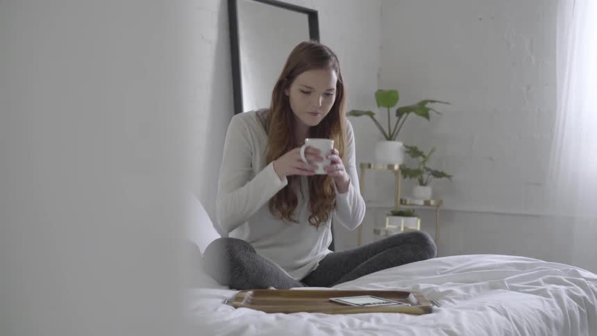 Woman sitting on bed drinking coffee while scrolling on her phone