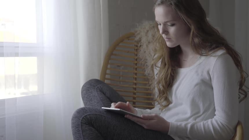 A teenage girl scrolls on a tablet while sitting next to a window with white curtains