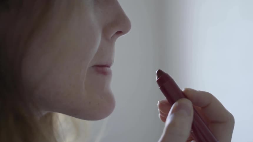 A white female uses her right hand to apply a mauve color lipstick pencil to her lips