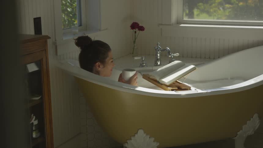 A white woman holds a cup and reads a book while sitting in the bathtub