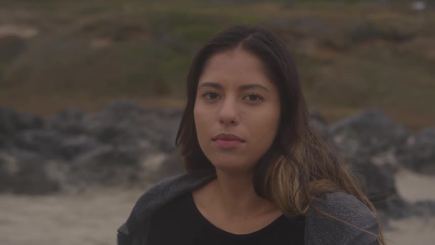 A woman is standing in front of a beach and hillside looking into the camera