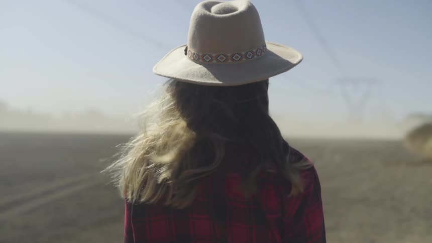 A woman in a faux cowboy hat stands still and looks out over a windswept stretch of open land in a dry and windy locale