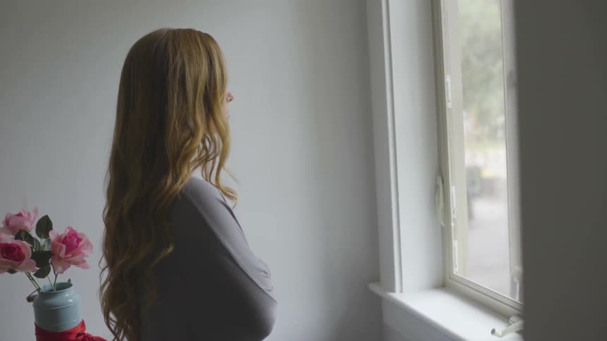 A white woman with crossed arms stares out of a window