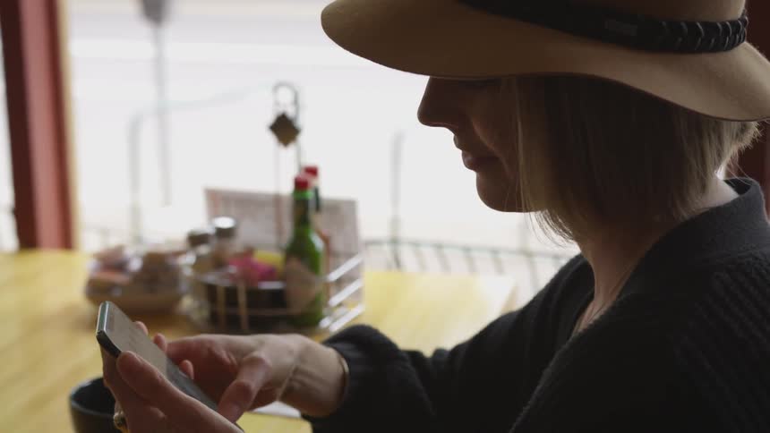 White woman sitting on a table on a restaurant next to the window using her cell phone