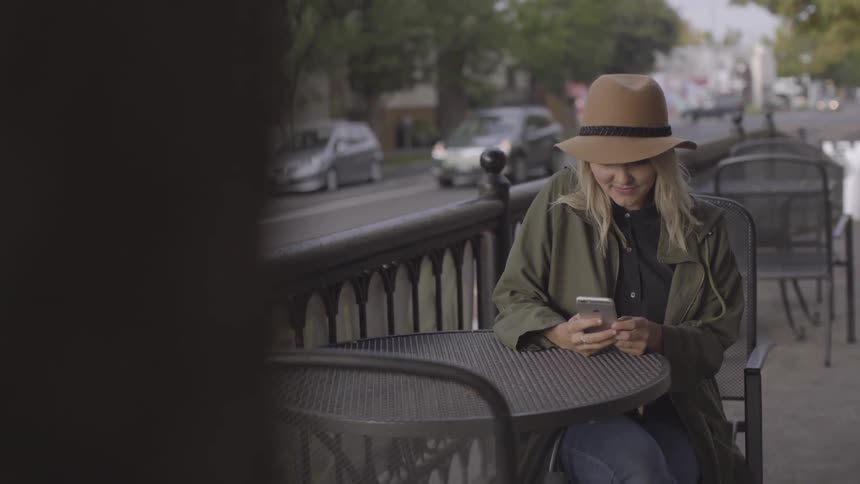 Women sits at the cafe table and checks her mobile phone while cars pass by