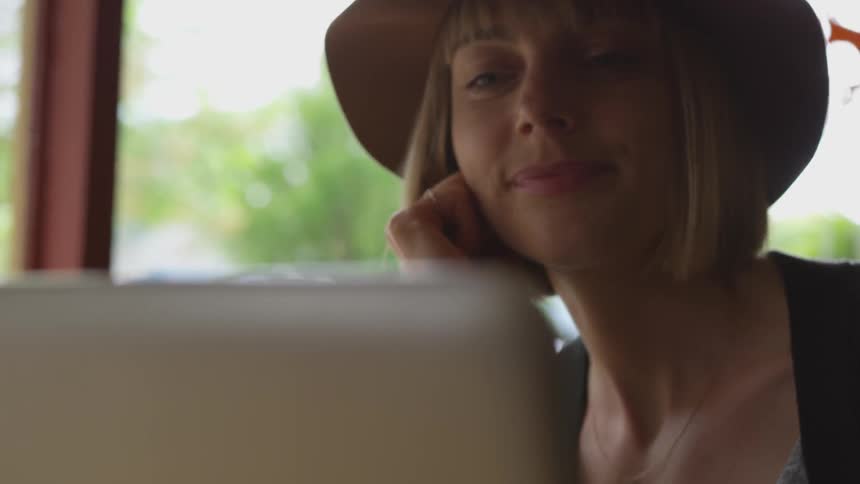 Girl wearing wide hat puts hand on chin and looks at laptop happily indoors during daylight