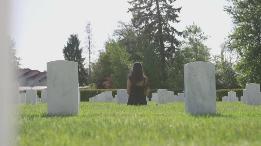 A woman dressed in black kneels in front of a tombstone in a cemetery