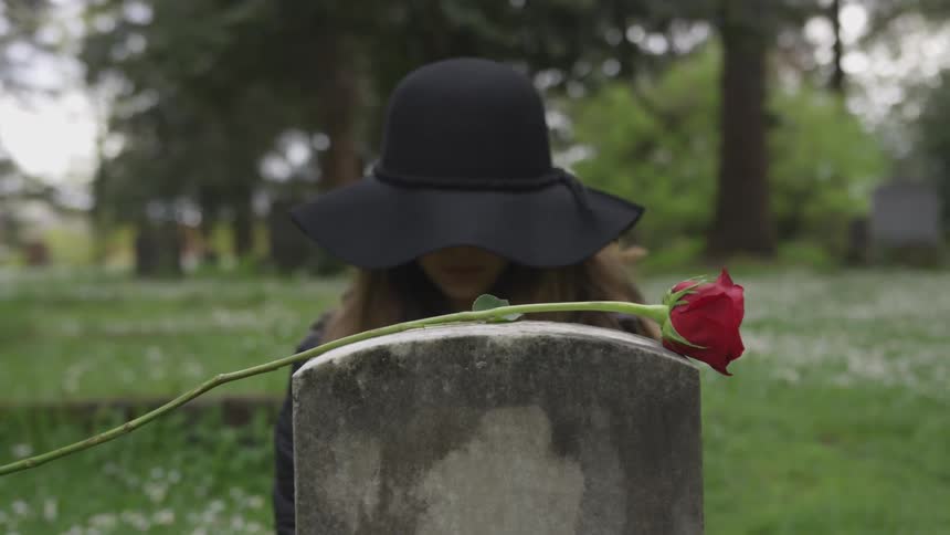 A woman sits in front of a headstone with a red rose in a cemetery