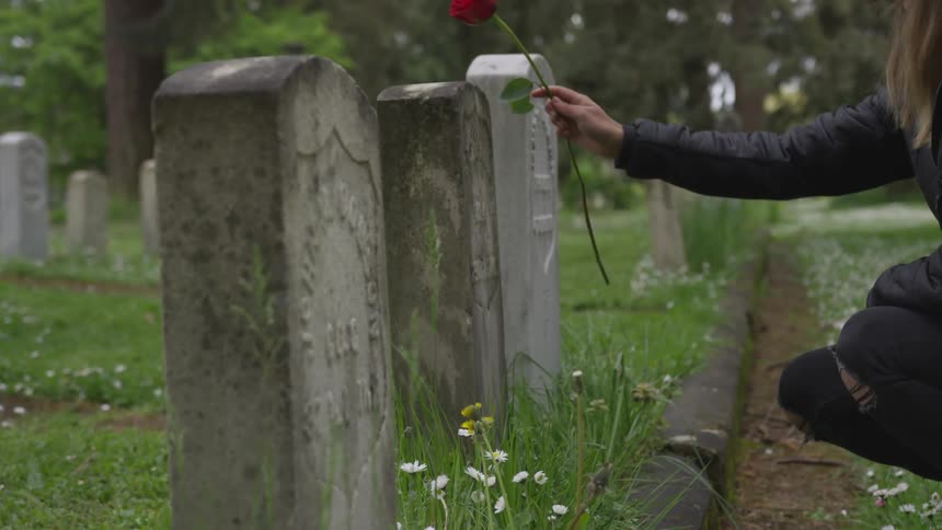 Blonde woman who wears black wide hat visits graveyard and puts a rose on a tombstone on a cloudy day