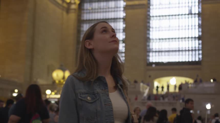 A young woman with a purse stands in the middle of the concourse in grand central station as she spins around to look up at every angle