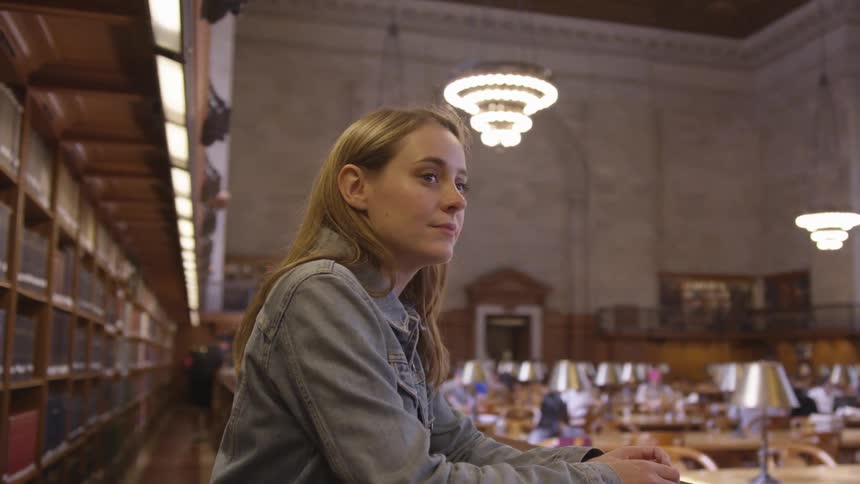 A woman leans on a railing overlooking the tables and chairs in a large library