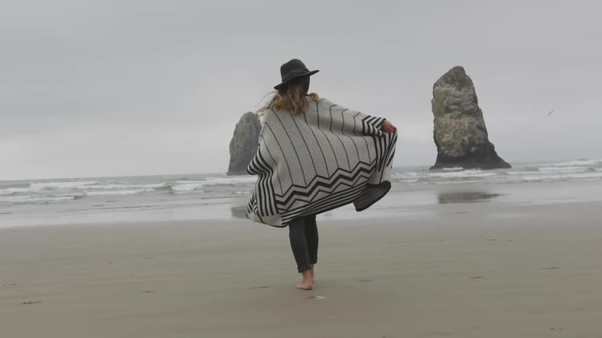 A woman wearing a poncho and hat walks on a beach