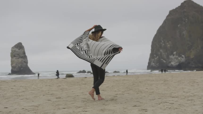 A white woman wearing a shawl and a hat walks on a windy beach