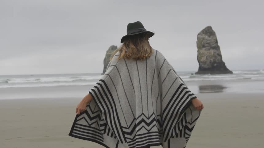 A woman in a black and gray poncho sweater and black hat walks on the beach towards the ocean