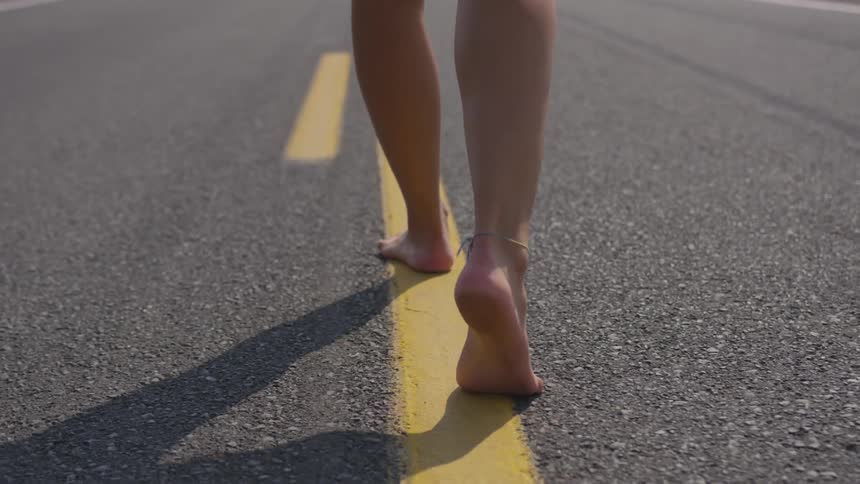 A barefoot woman wearing an anklet walks down the center of a two lane highway