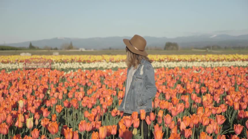 A woman with denim coat and a brown hat strolls through a field of tulips