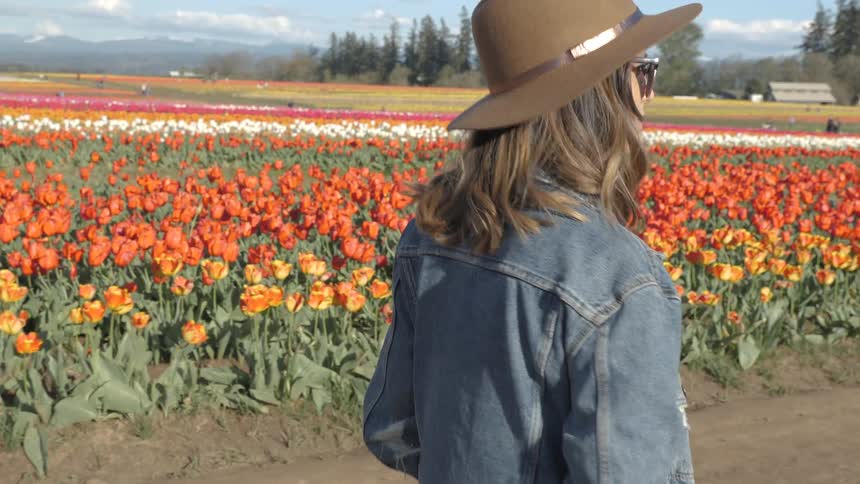 Woman in brown hat and denim jacket walking next to a field of tulips in the country