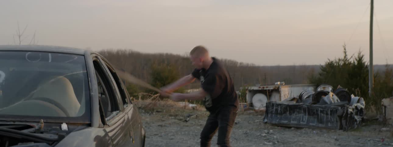 Boy hits junk car with baseball bat furiously at dusk