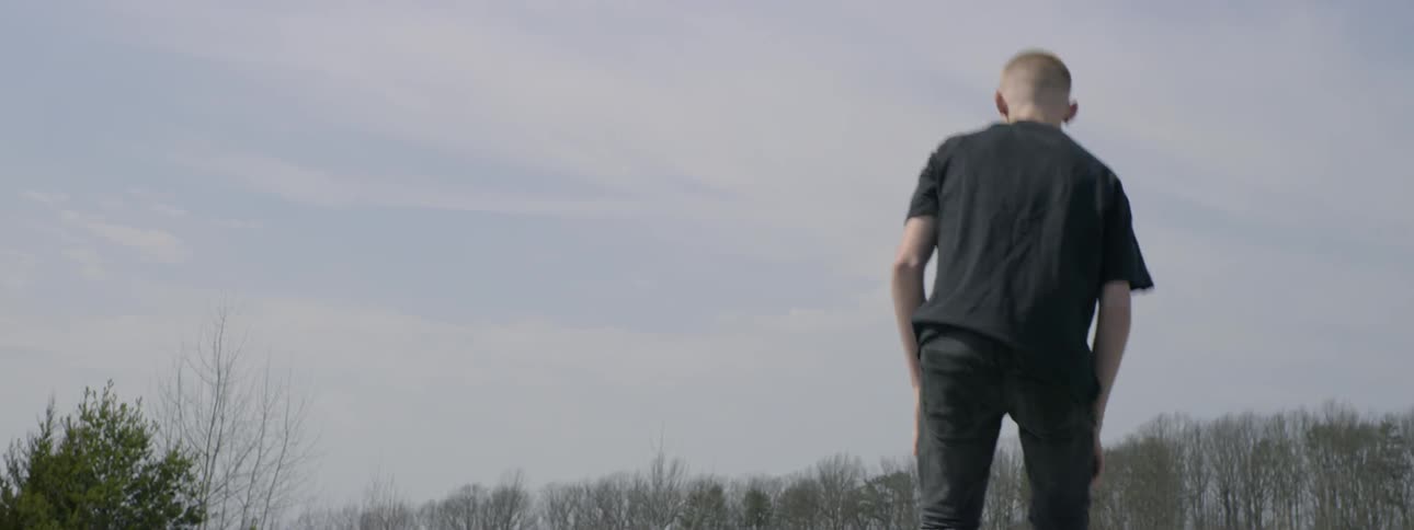 A teenager jumps up on the top of an old car at the junkyard and then kneels on the roof