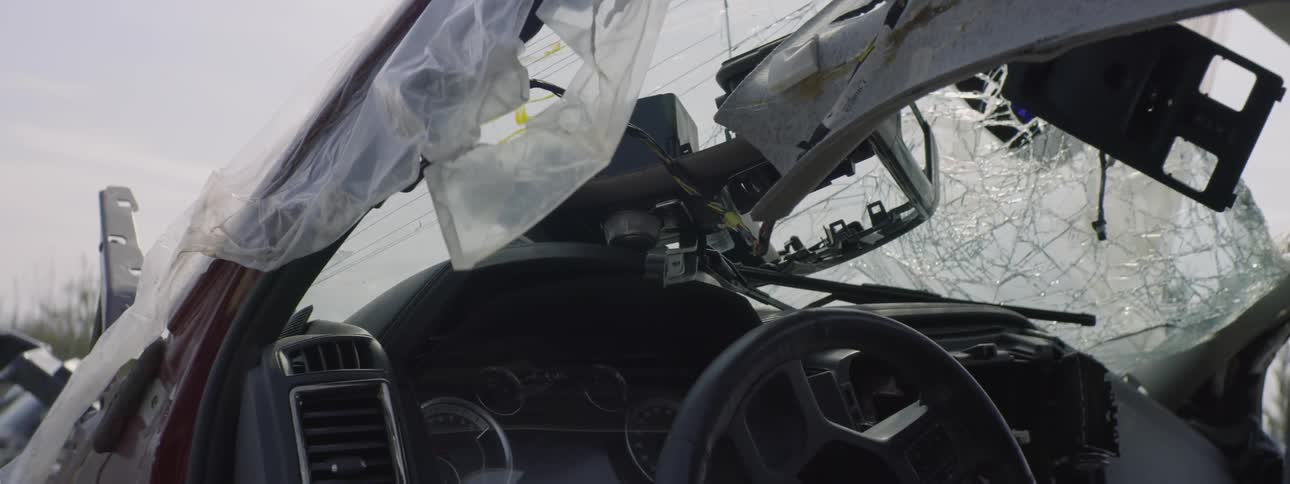 The remains of a wrecked car with a shattered window and bent frame sits draped in plastic as a breeze gently moves the plastic