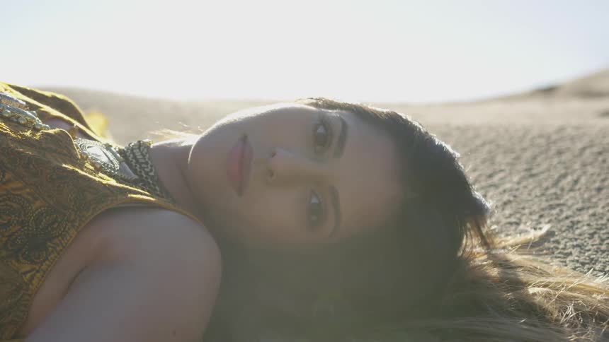 The woman in yellow with dark hair that is blond on the tips is laying on the desert floor slowly turns her head to look at the camera