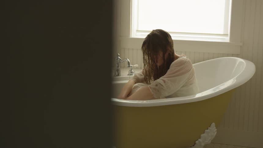 A woman with damp hair hanging in front of her face is sitting hunched forward in a bathtub in a well lit bathroom