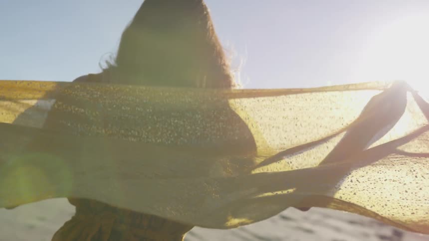 A woman walks along the beach holding a shawl over her shoulders