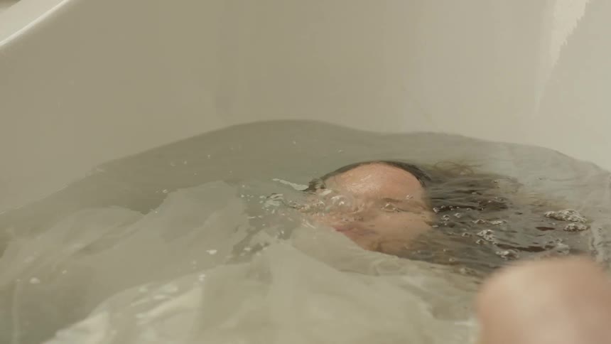 A white woman submerges herself underneath the water in a white bathtub