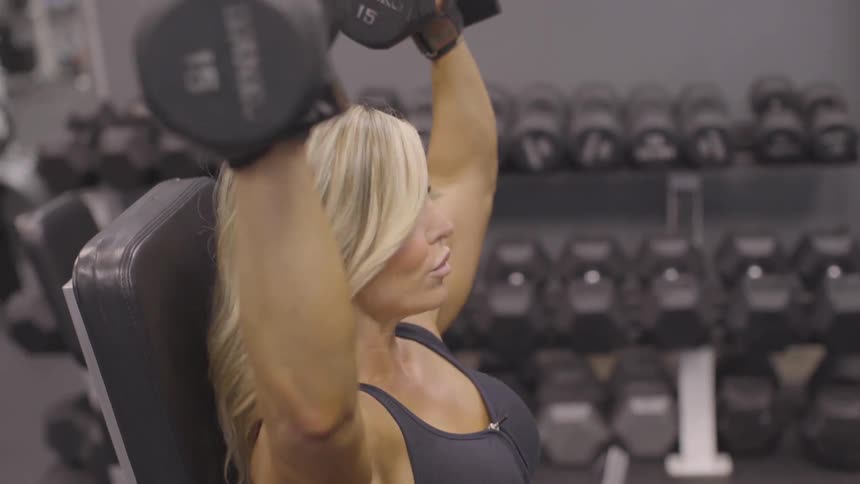 A blond woman is doing overhead shoulder presses with 15 pound free weights in a gym