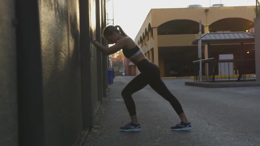 A white woman in workout clothing stretches against a building