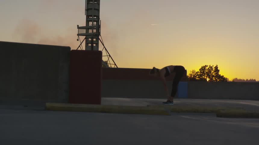 A young white woman wearing yoga pants and a sports bra stretches outside