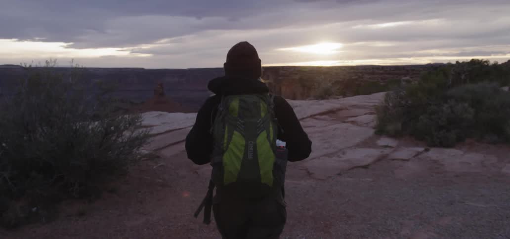 A hiker wearing a backpack walks toward the edge of a cliff at sunrise