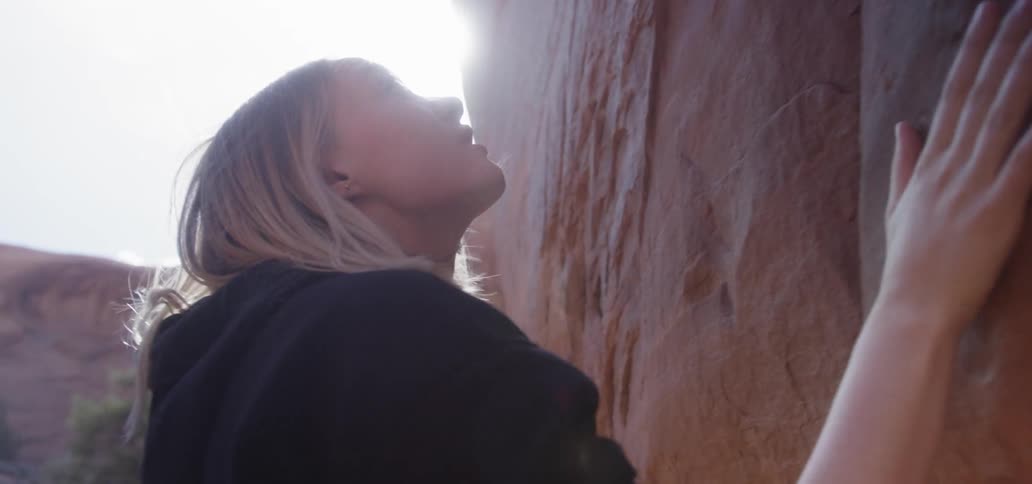 A blonde haired woman touching a red boulder outside