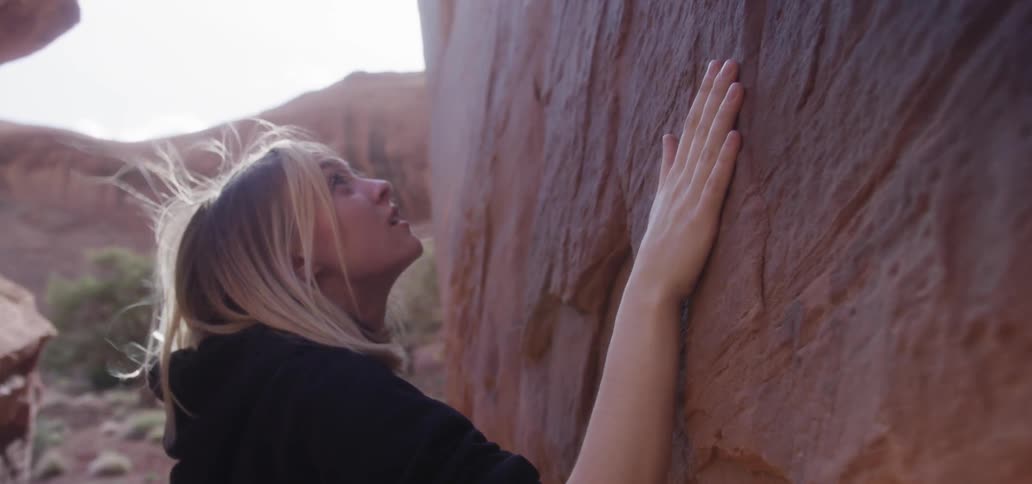 Woman slides her hand along the side of a large rock as the breeze blows her hair