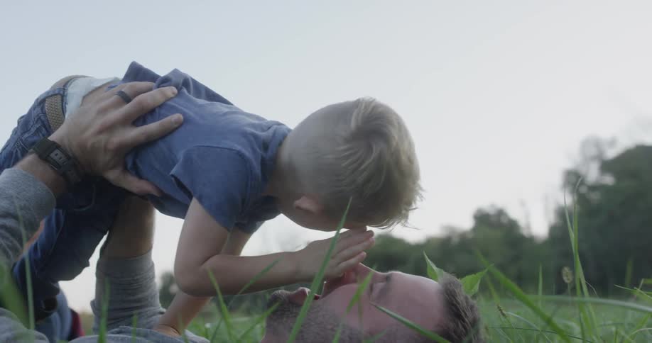 The boys daddy is laying on the grass he laughs as his father lift him up above his face and brings him down so they could touch noses