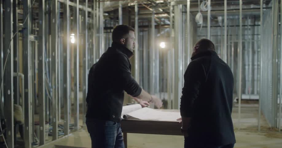 Three men in an unfinished structure are examining blueprints on a table