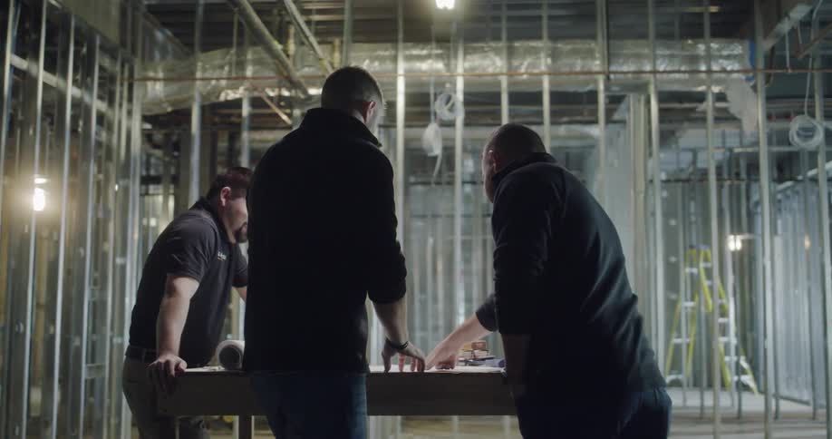 Three men look at building plans laying on a desk in a construction site
