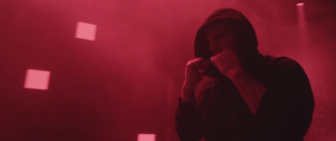 A man wearing a hoodie with the hood up shadow boxes while red lights flash and blink on a wall