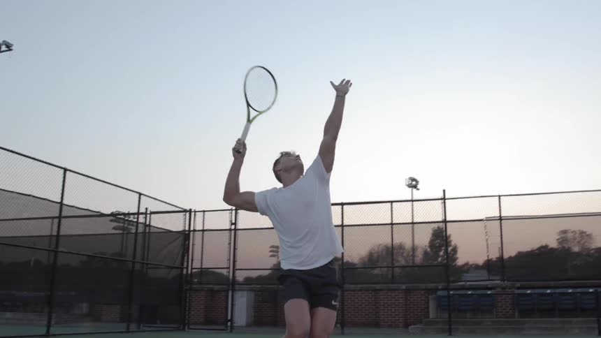 A man hits a serve on an outdoor tennis court as the sun sets behind him