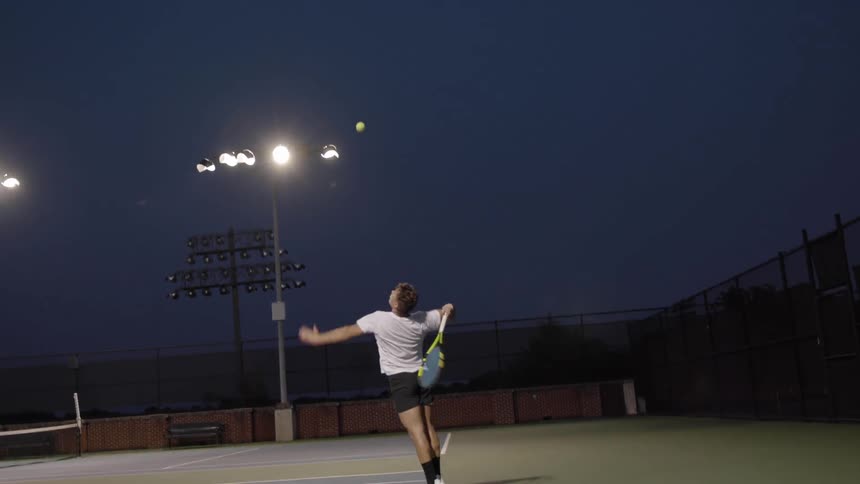 A white male throws a tennis ball straight up to serve across a tennis court illuminated by spotlights