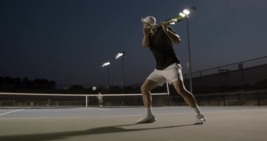 Two young men play tennis on a lighted court at night