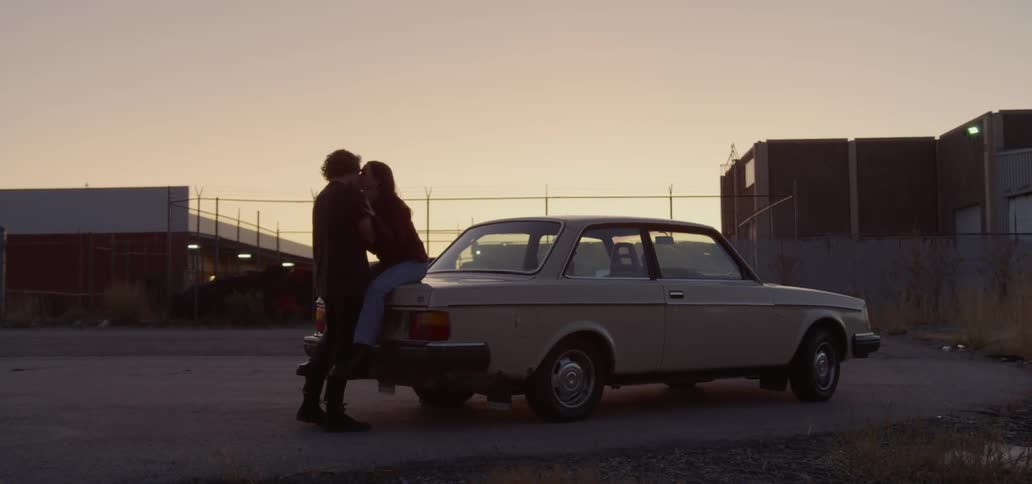 A man kisses his girlfriend who is sitting on the trunk of their car in a parking lot at sunset