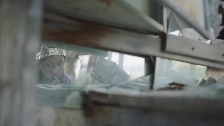 A girl wearing a crown puts lipstick on while looking in cracked and jagged pieces of mirror