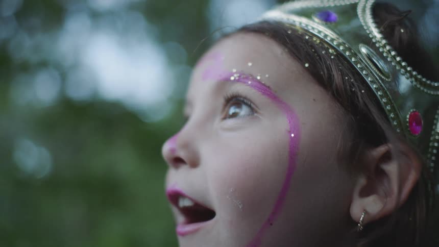 A young girl with purple paint on her face and a crown on her head looks up in wonder and smiles